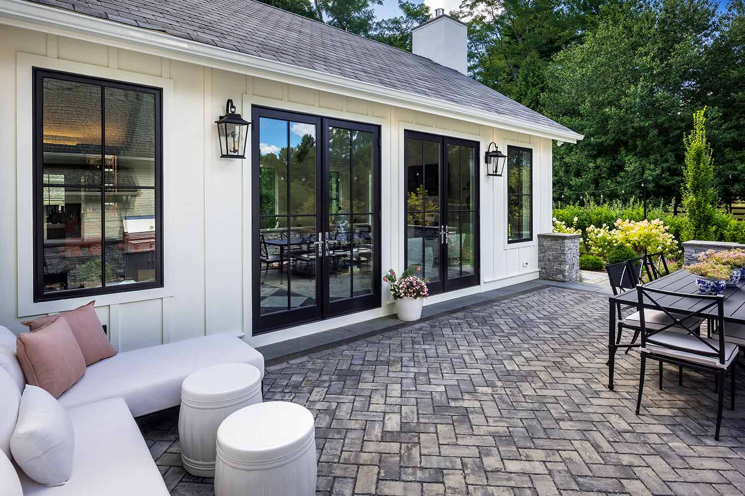 A modern white home exterior with black-framed glass doors and windows opening onto a patio with paver flooring, outdoor furniture, and landscaping.