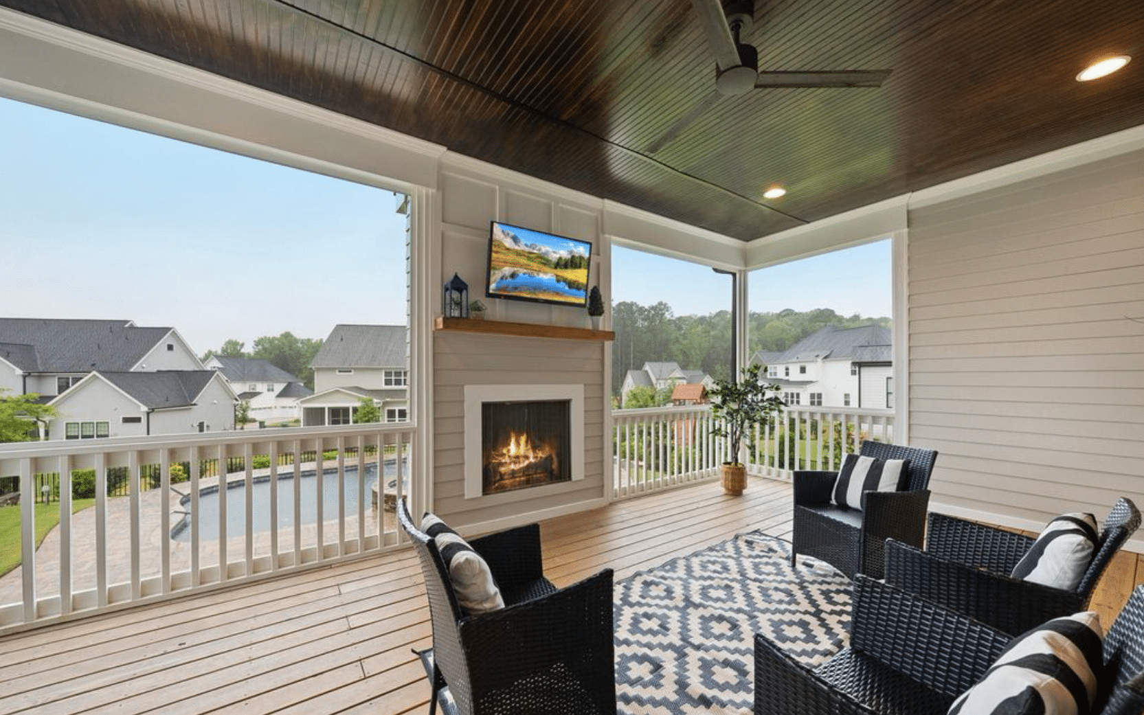 A covered deck with composite decking, black wicker furniture, a fireplace with TV mounted above, white railings, and views of a residential neighborhood.