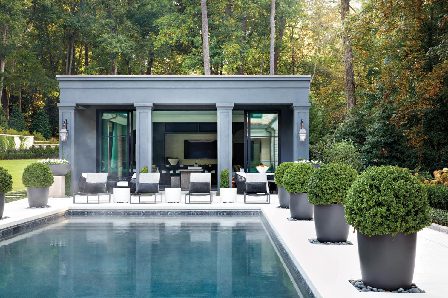 A modern residential pool house with gray exterior, large glass doors opening to an interior lounge area, surrounded by white pool deck, manicured landscaping, and forest backdrop.