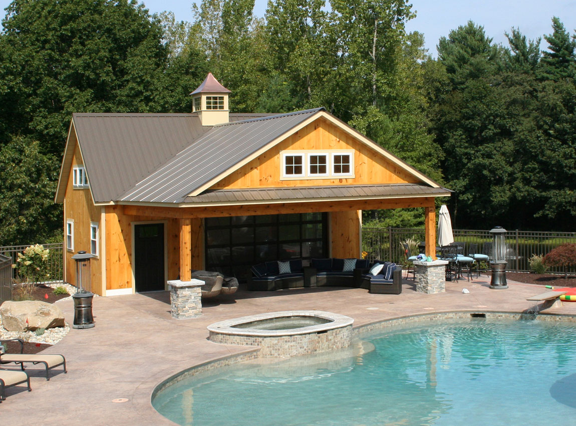 A modern yellow wooden pool house with metal roof and cupola stands beside a backyard swimming pool with hot tub, surrounded by a brick patio and landscaping.
