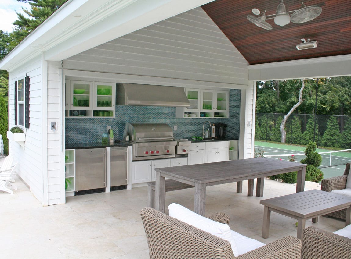 An outdoor covered patio kitchen with white cabinetry, stainless steel appliances, teal tile backsplash, wooden dining table, and views to a tennis court and landscaped yard.