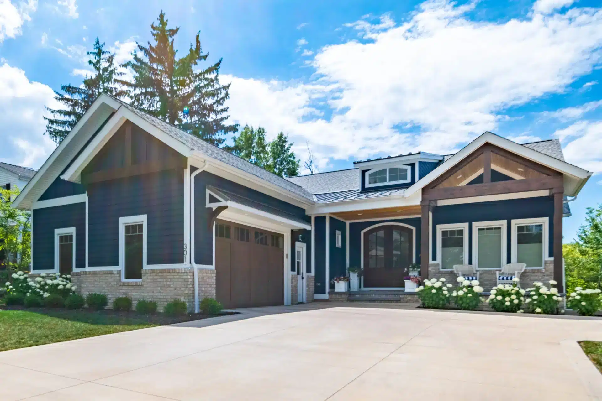 A modern two-story home with dark blue exterior siding, brick accents, multiple garage doors, covered entry porch, and mature evergreen trees in the landscaping.