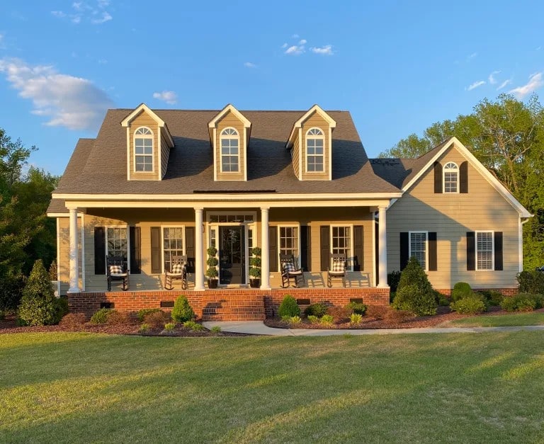 A yellow and white two-story residential home with three dormers, brick foundation, front porch with columns, and manicured landscaping on a sunny day.