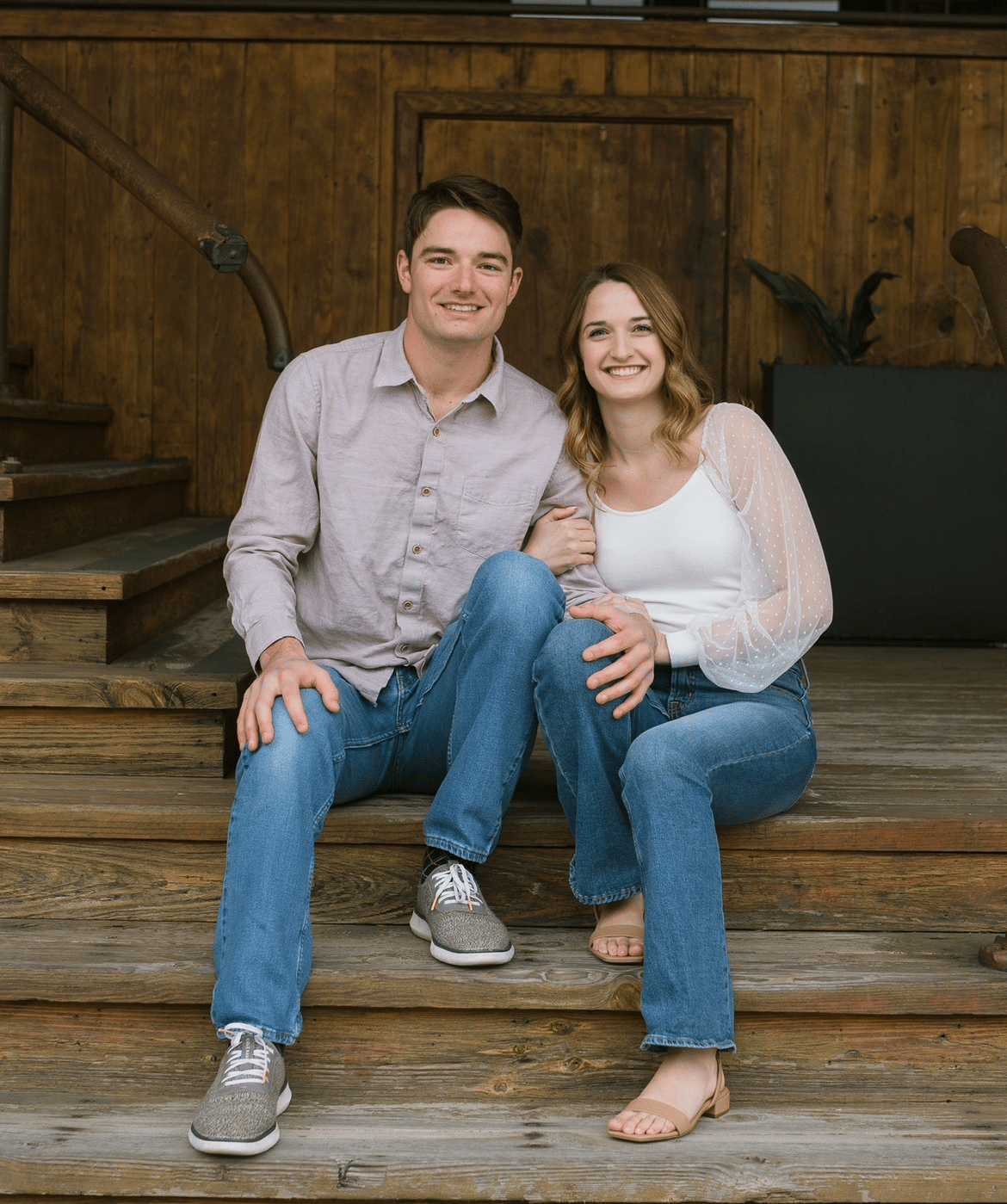 Two people smiling while sitting together on wooden steps in front of a wooden wall with architectural details.