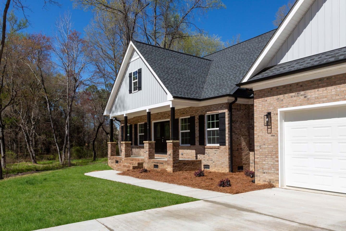 A newly constructed brick home with a covered front porch, white trim, gabled roof, and attached garage, situated on a grassy lot with bare trees and landscaping.