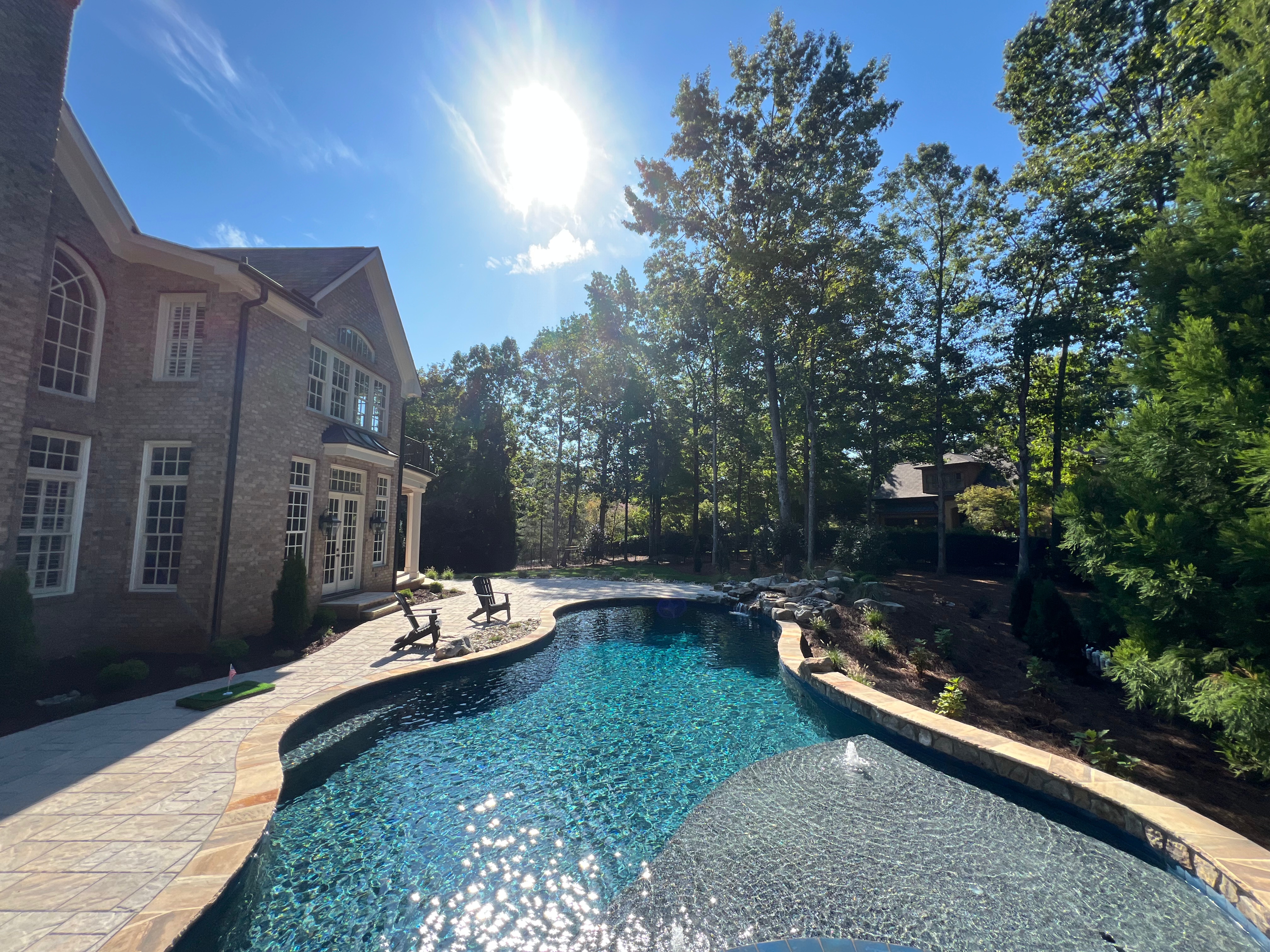 A luxury residential backyard features a curved swimming pool with blue water, surrounding paver patio, Adirondack chairs, brick home, and mature trees in a wooded setting.