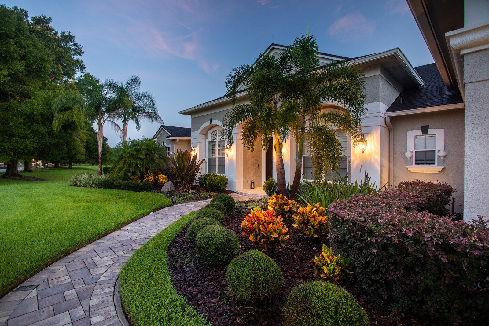 A modern single-story home at dusk with stucco and stone exterior, illuminated landscape lighting, manicured lawn, curved paver walkway, and tropical plantings including palm trees and flowering shrub